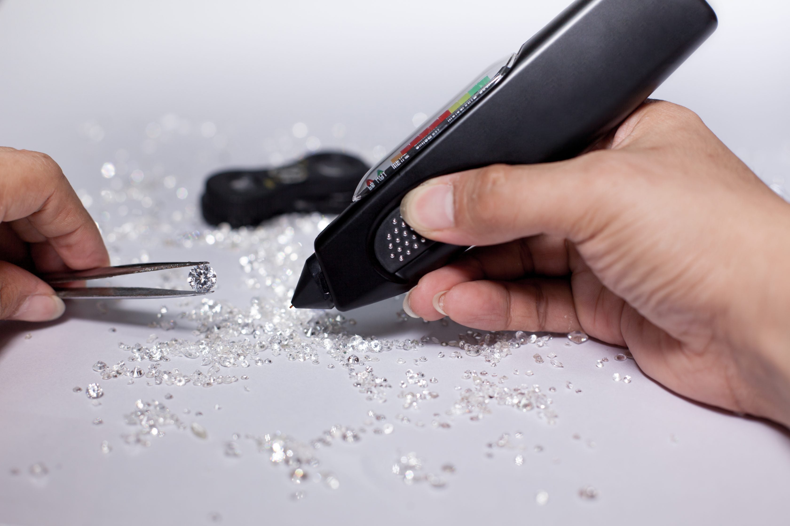 Diamonds on tweezers with testing machine on a white background, reflections on the ground. brilliant cut diamond held by right hand, tester, white gemstone, diamond checking equipment.